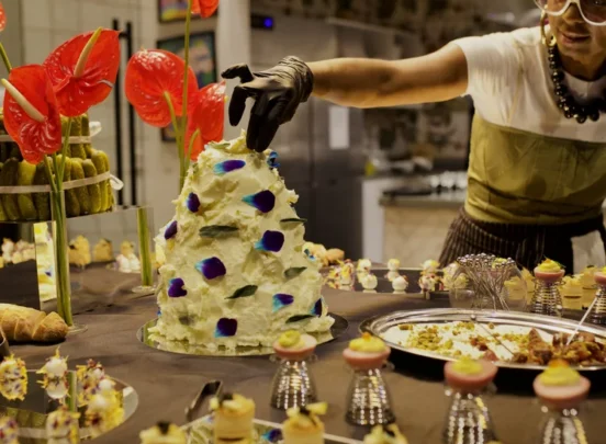 A chef decorates a tall, white cake with blue flowers, surrounded by assorted desserts and red flower arrangements in a bakery kitchen.
