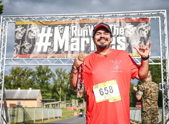 A smiling runner in a red shirt holds a medal at a race finish line, with a #RunWithTheMarines banner in the background.