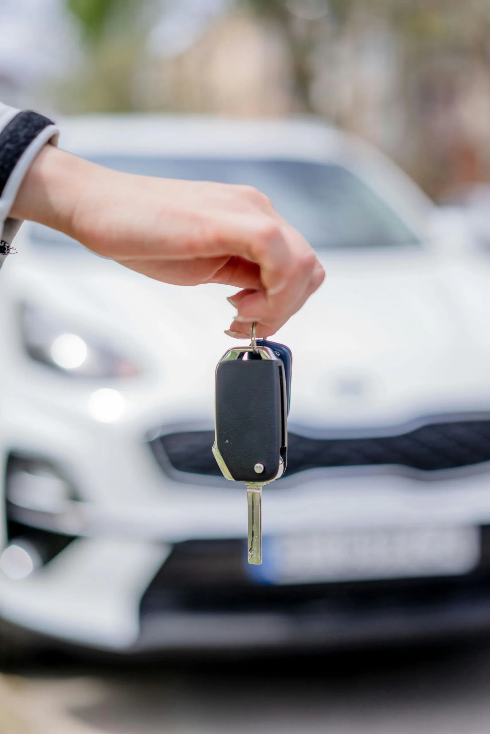 A hand holding a car key in front of a white car, with the vehicle slightly out of focus in the background.