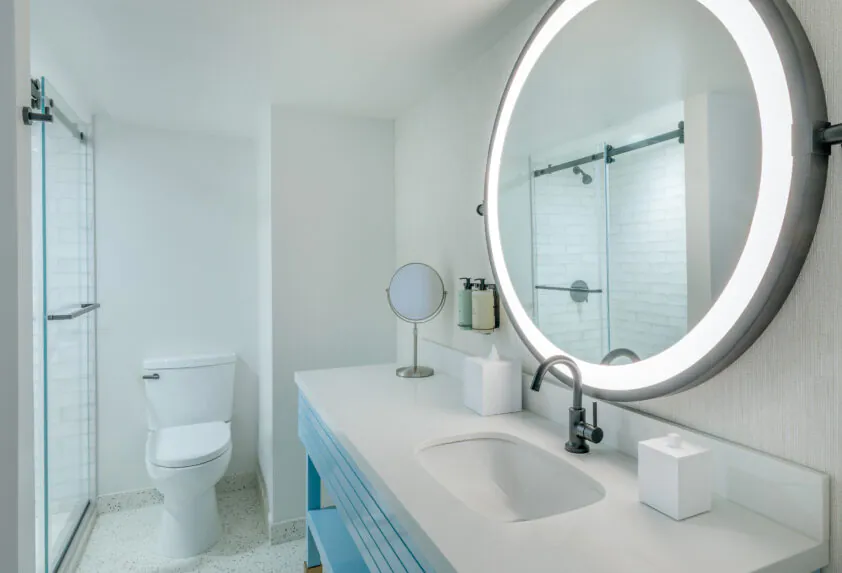 Modern bathroom with a large round backlit mirror, white sink, blue vanity, toilet, and glass shower enclosure in a bright, minimalist style.