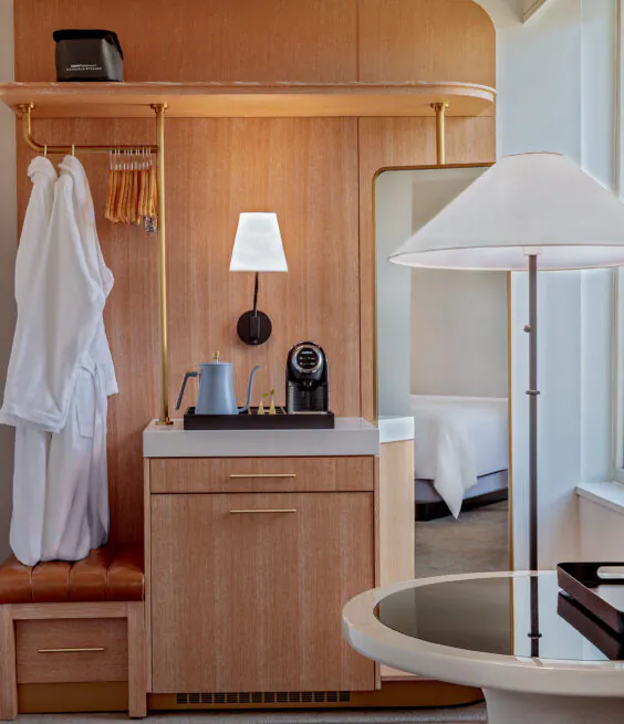 A hotel room corner with a robe on a hook, a coffee maker on a wooden cabinet, a round table with an ice bucket, and a floor lamp nearby.