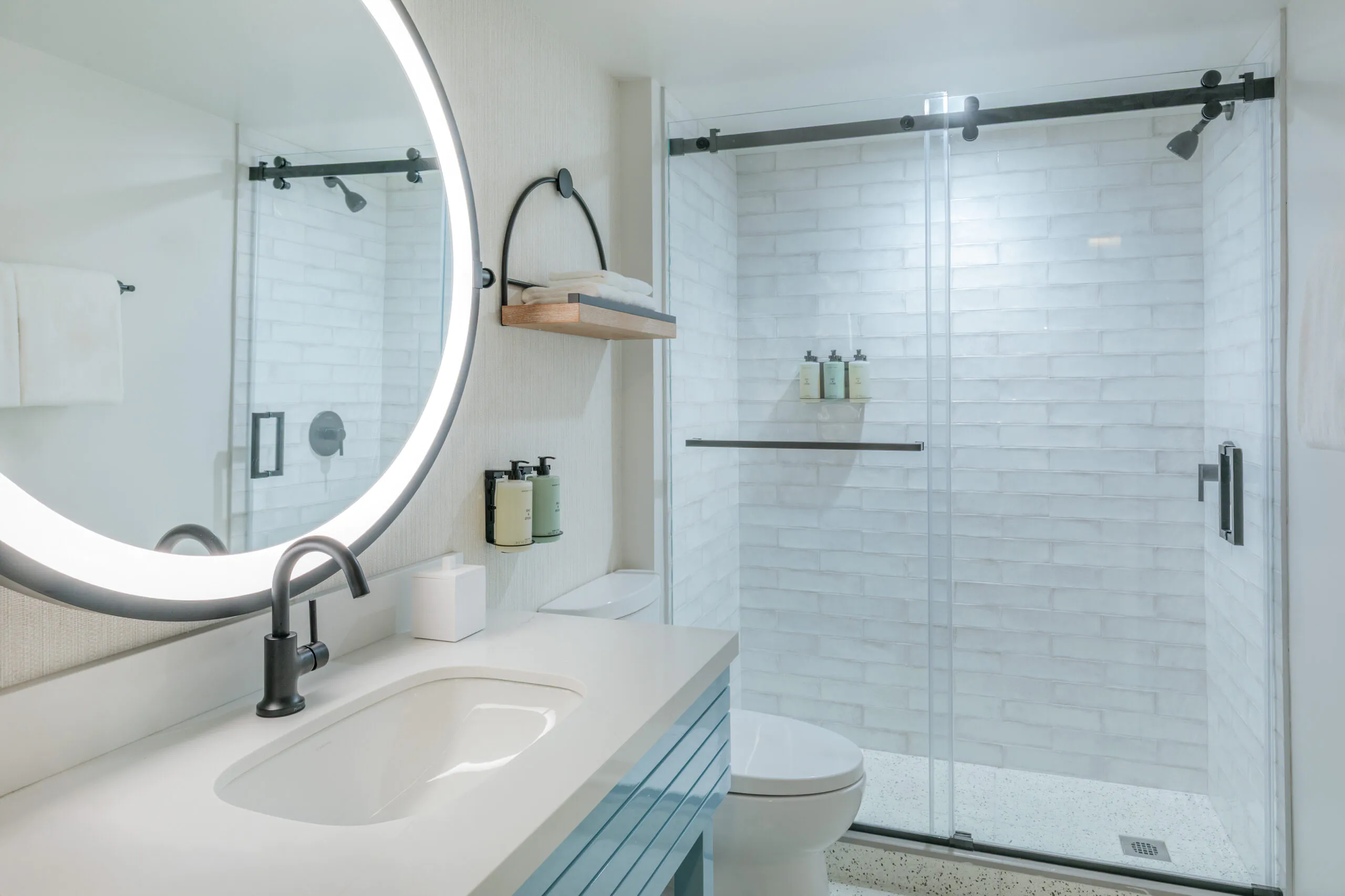 Modern bathroom with a glowing round mirror, white sink, wall-mounted soap, and a glass shower with white subway tiles and bottles on a shelf.