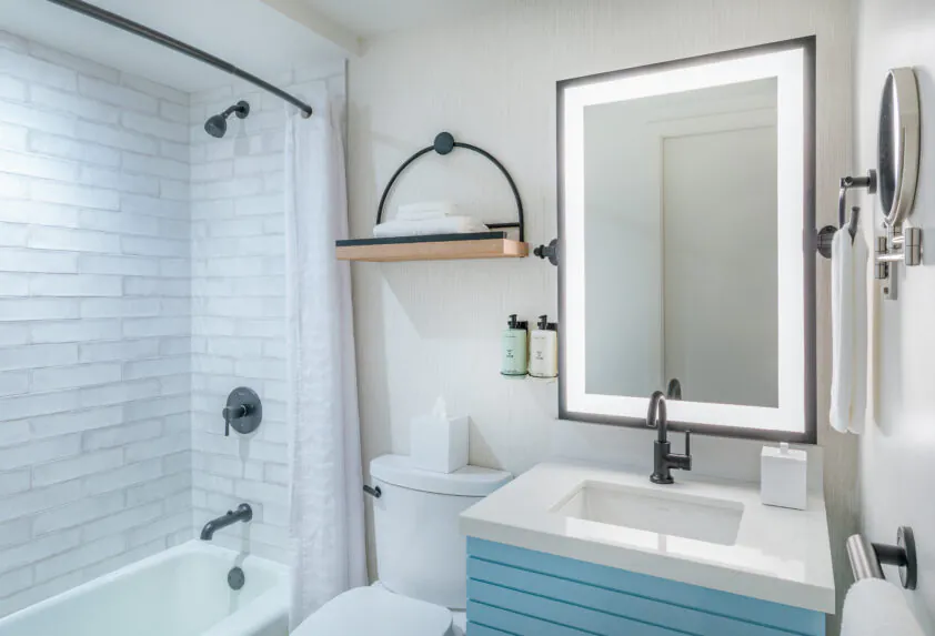 Modern bathroom with a lit mirror, white sink, toilet, bathtub with white tile, wall shelf, and neatly arranged toiletries and towels.