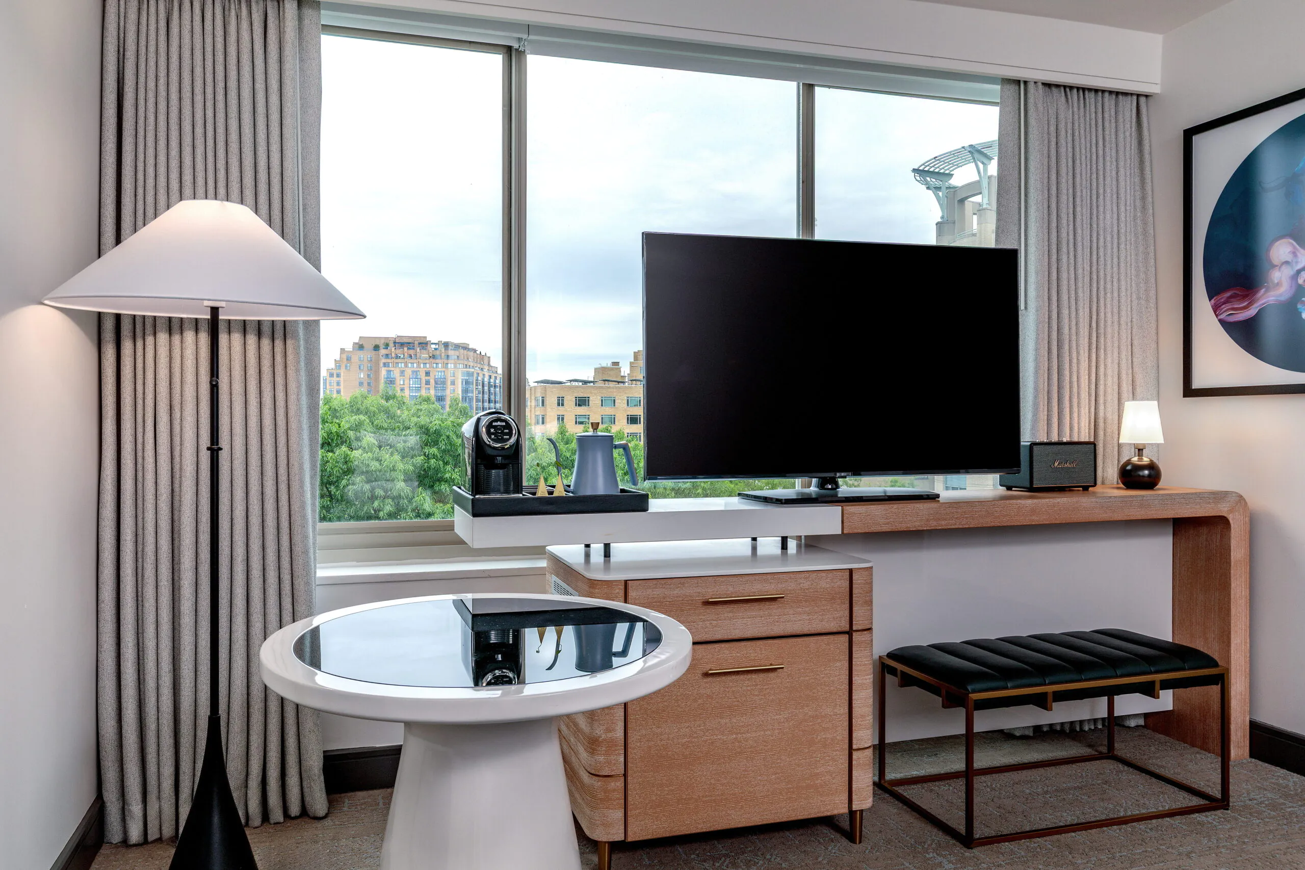 Modern hotel room corner with a TV on a wooden desk, a round table, a floor lamp, and large window with city and greenery view.