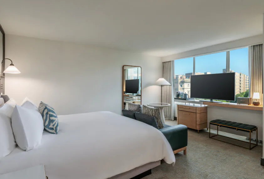 Modern hotel room with a large bed, TV, desk, bench, and window showing a city view under a clear blue sky.