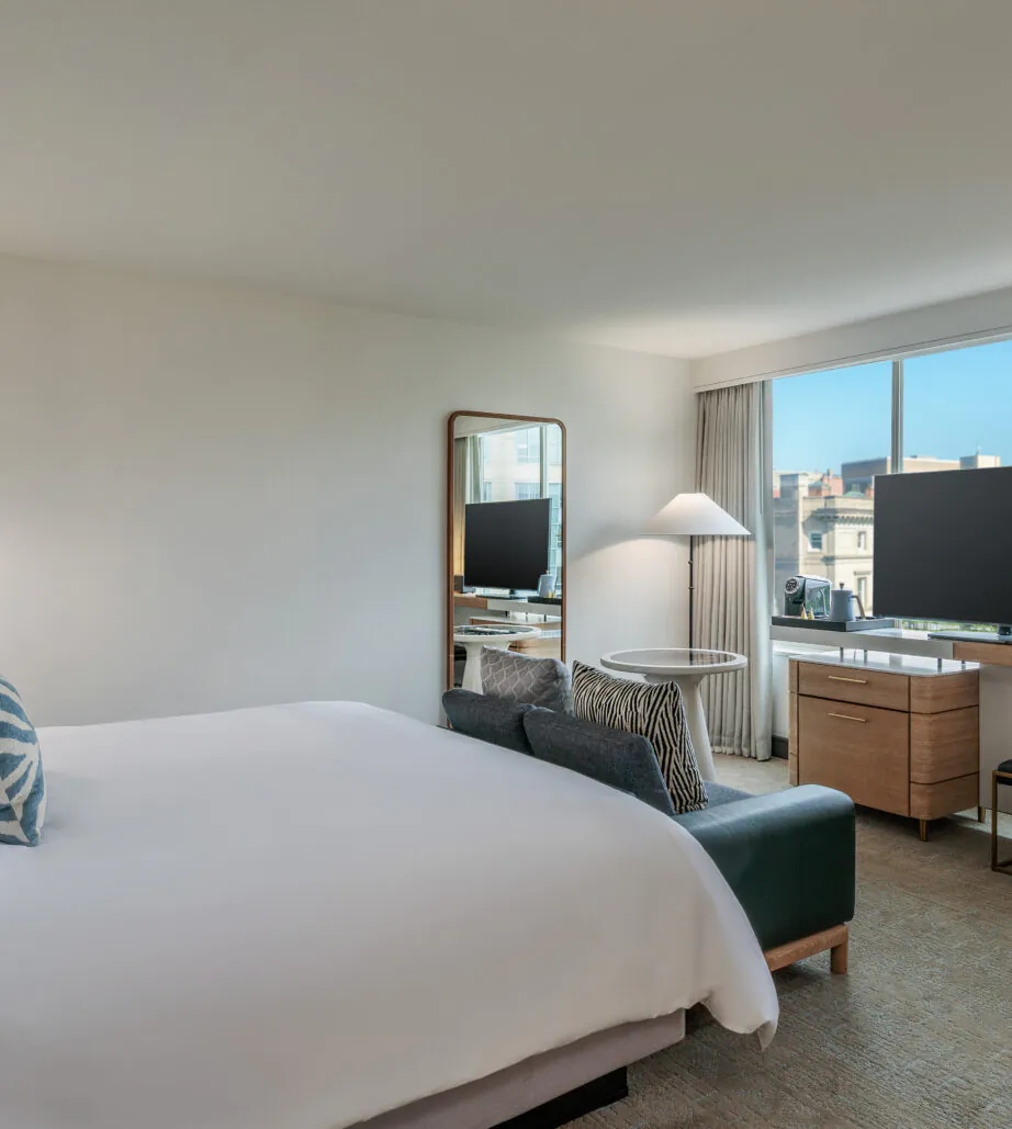 Modern hotel room with a large bed, TV, desk, bench, and window showing a city view under a clear blue sky.