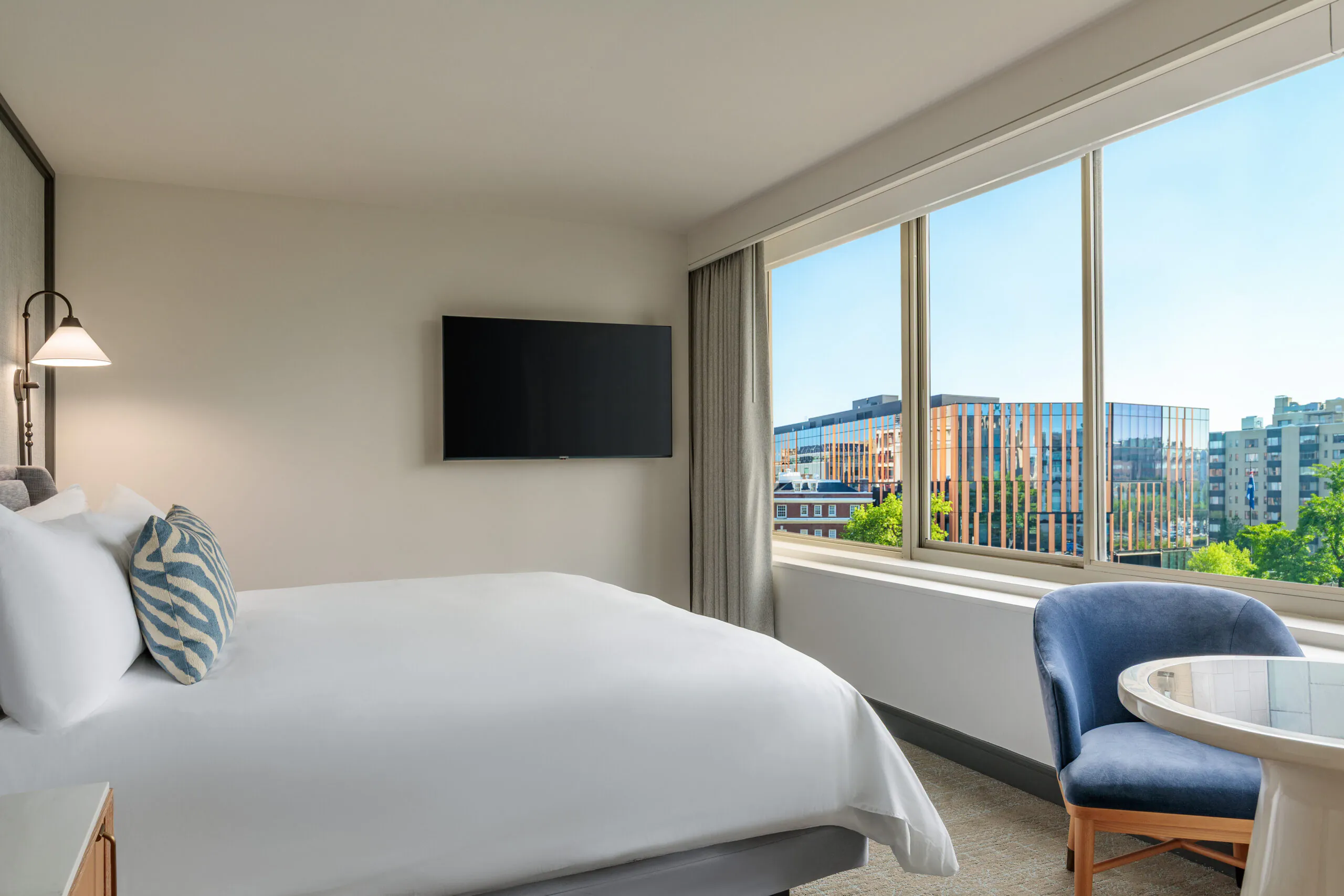 A hotel room with a neatly made bed, wall-mounted TV, blue chair, round table, and large window overlooking modern city buildings.
