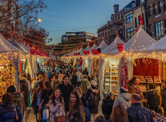 Crowd of people walking through a festive outdoor holiday market at dusk with white tents, string lights, and decorated stalls.