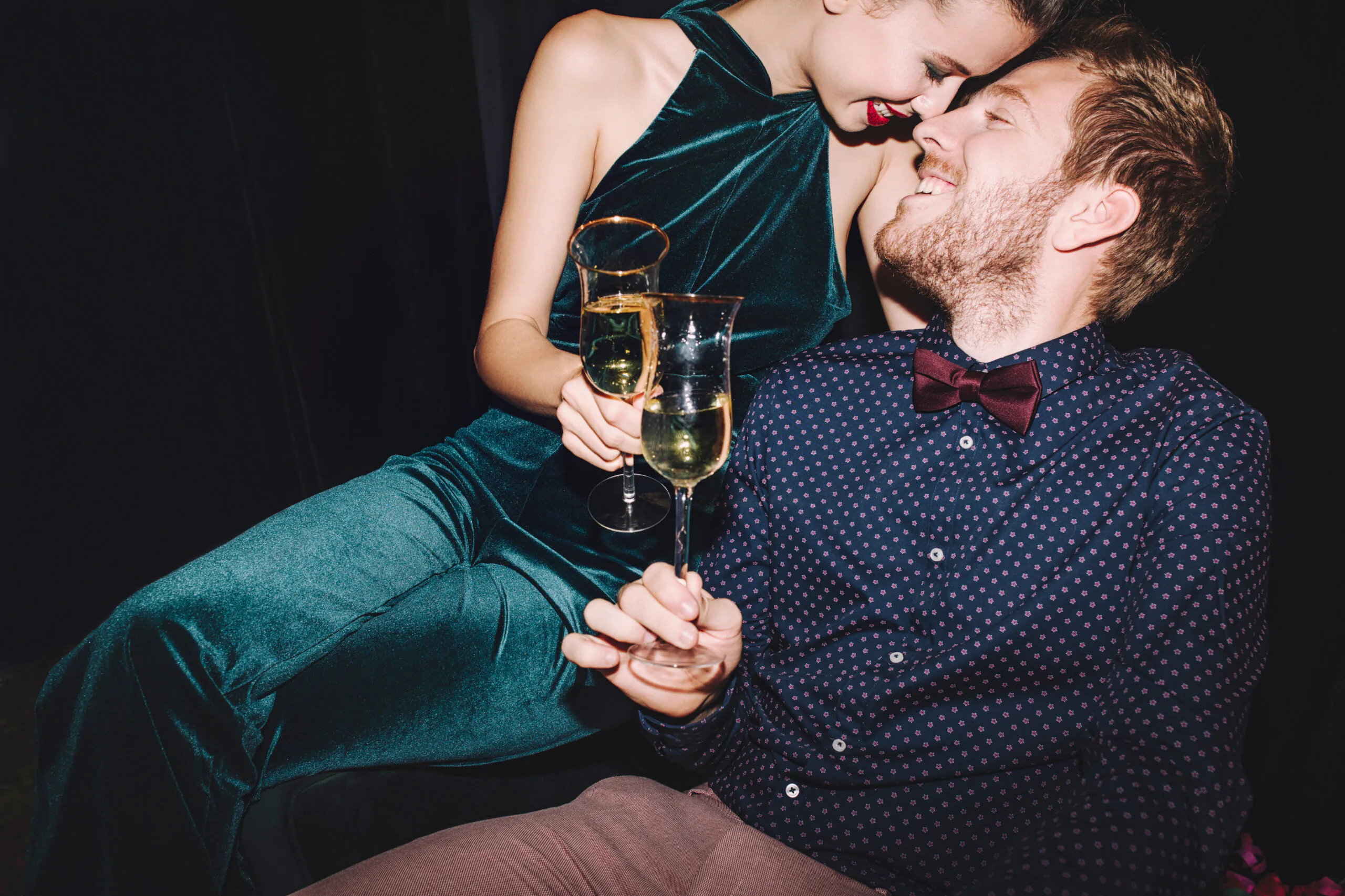A smiling couple in formal attire sit close together, holding champagne glasses and toasting at a celebration or party.