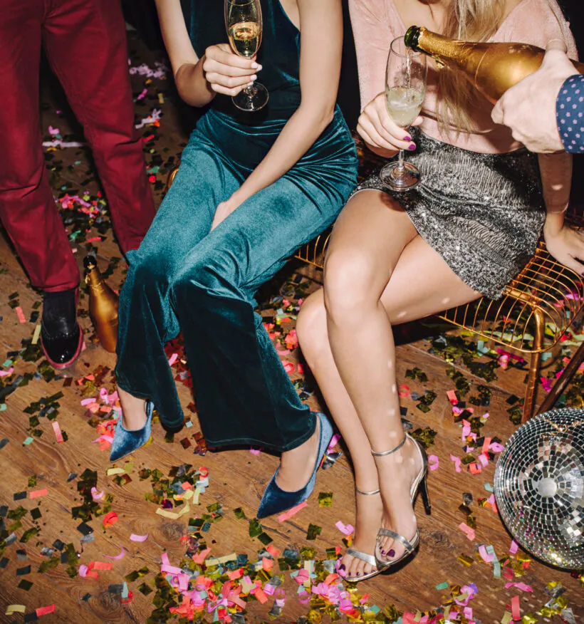Four people at a party, two seated with drinks, one pouring champagne, confetti on the floor, and a disco ball nearby. Festive atmosphere.