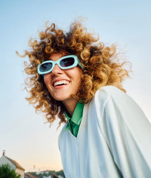 Smiling woman with curly hair and blue sunglasses stands outdoors, wearing a white sweater. Houses and a clear blue sky are in the background.