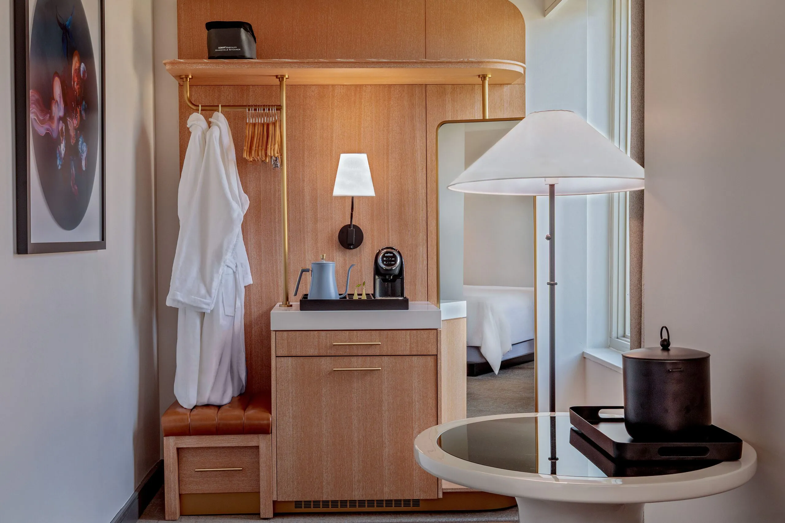 A hotel room corner with a robe on a hook, coffee maker on a wooden cabinet, table with a lamp, and an ice bucket, near a bed and a window.