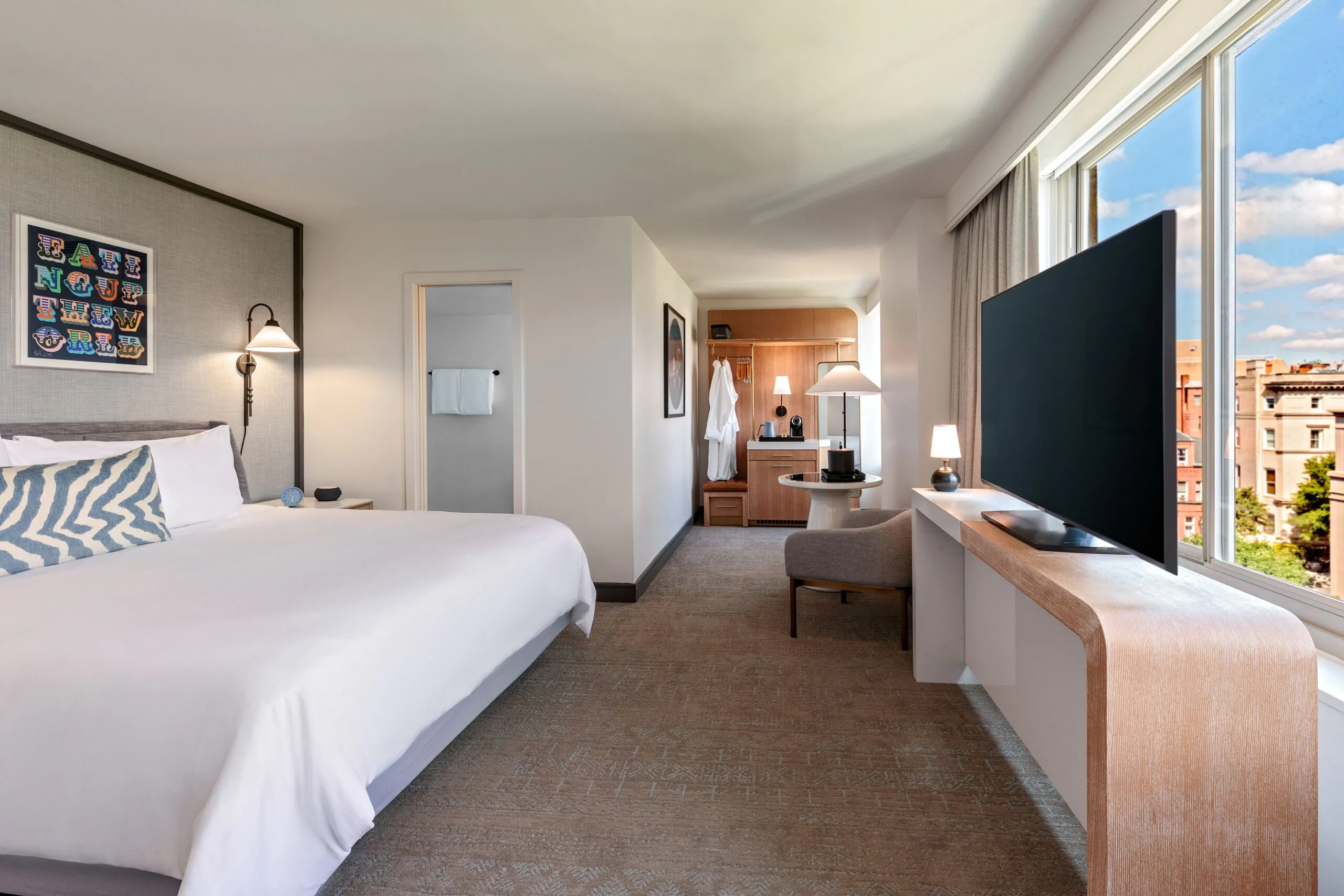 Modern hotel room with a large bed, TV, desk, armchair, closet area, and a window with a city view under a blue sky with clouds.