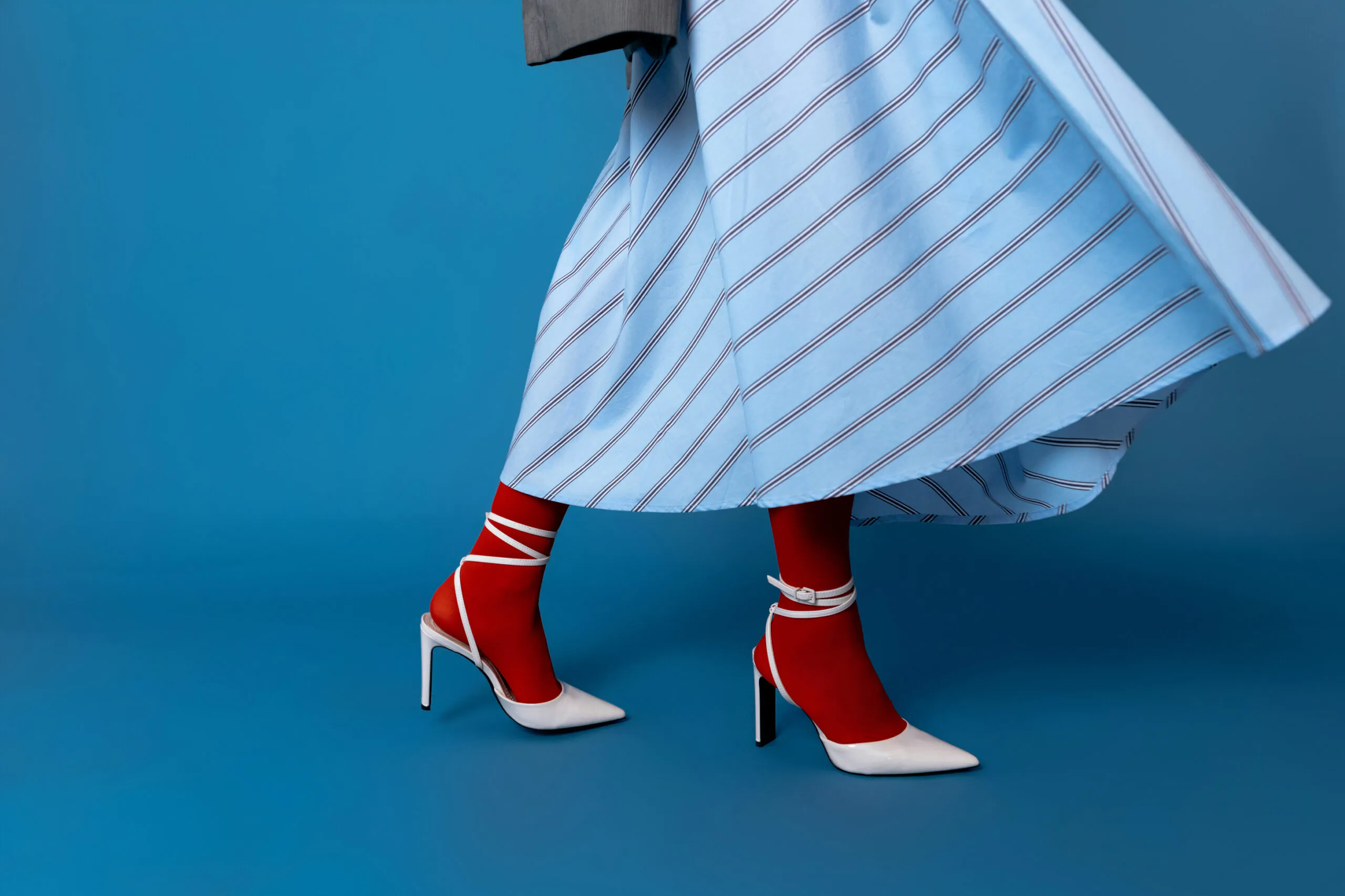 Person wearing a blue striped skirt, red tights, and white high-heeled shoes with ankle straps, standing against a solid blue background.