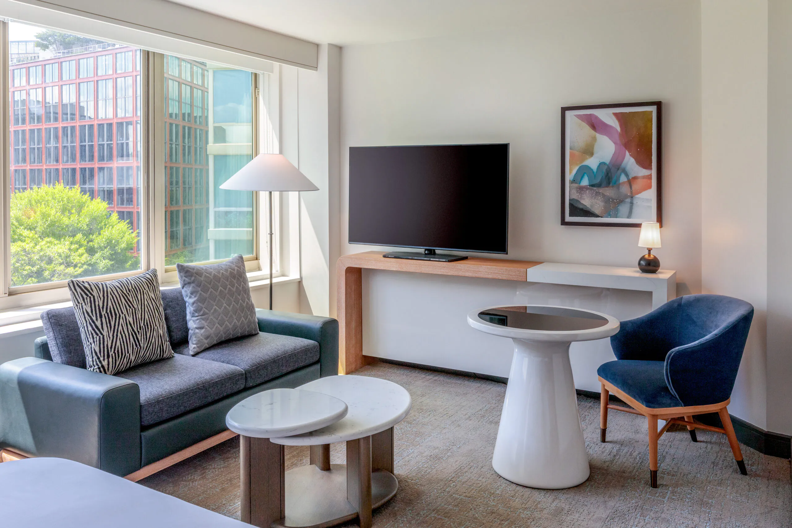 Modern hotel room with a sofa, blue chair, round tables, large TV, abstract art, and a window overlooking buildings and greenery.