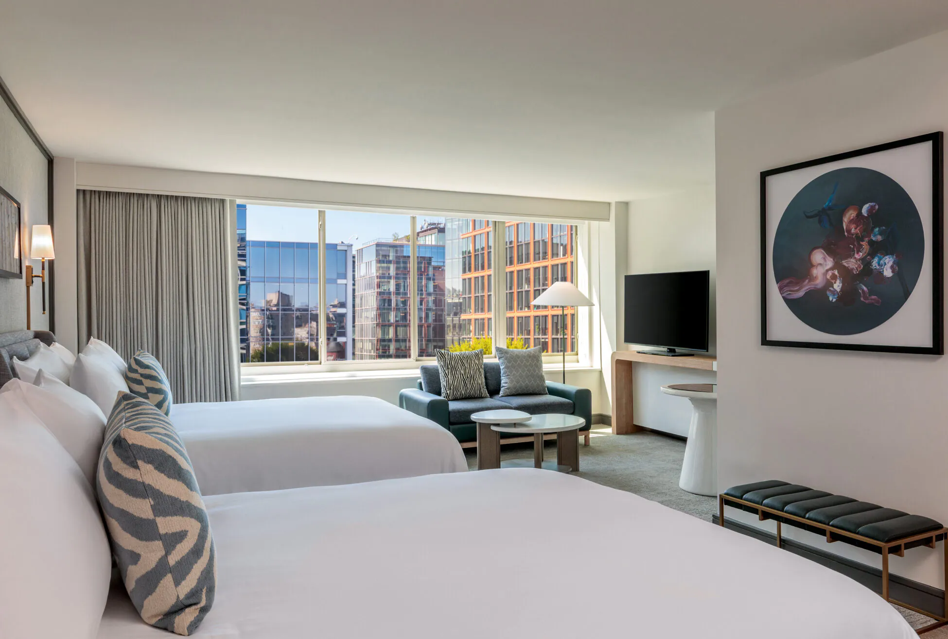 Modern hotel room with two beds, large window with city view, sofa, TV, round table, and decorative wall art above a bench.