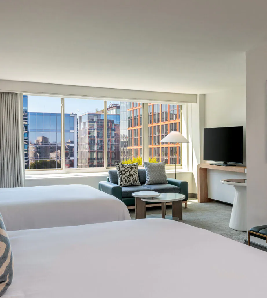 Modern hotel room with two beds, large window with city view, sofa, TV, round table, and decorative wall art above a bench.