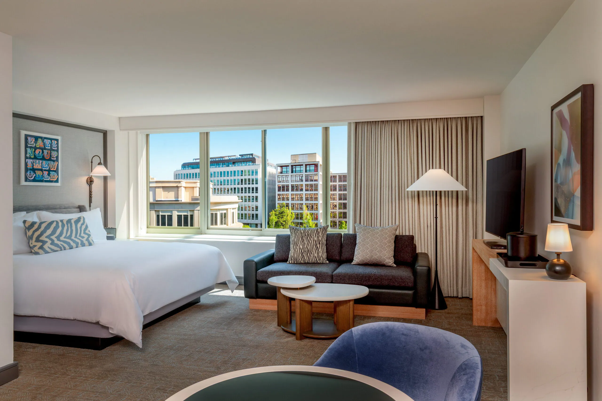 Modern hotel room with large bed, sofa, TV, coffee table, and city view through wide windows; neutral decor with blue and gray accents.