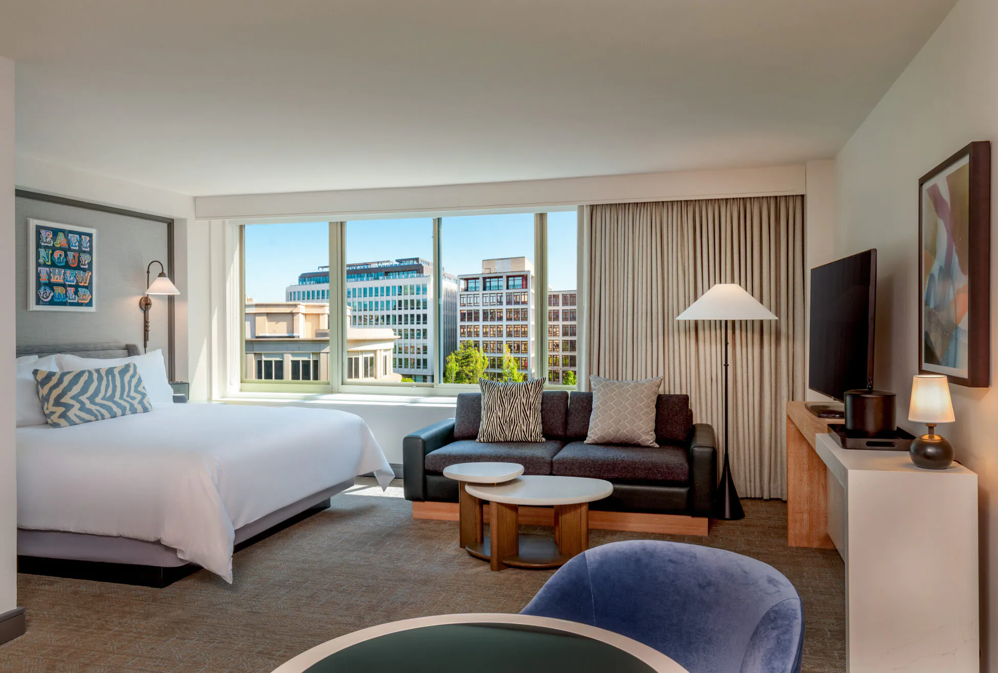 Modern hotel room with a large bed, sofa, TV, and coffee table; large windows show city buildings and blue sky outside.