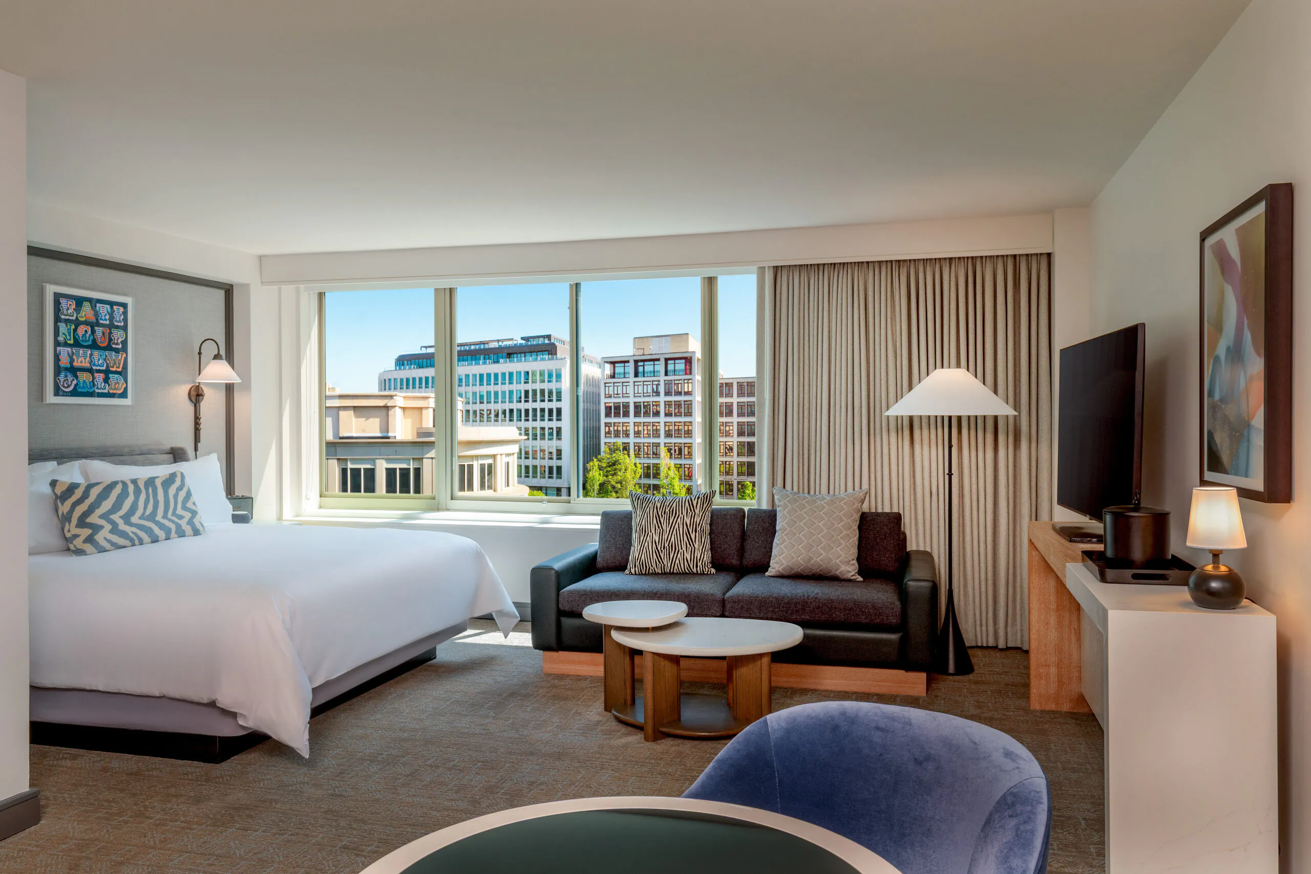 Modern hotel room with a bed, sofa, TV, coffee tables, large windows, and city buildings visible outside on a sunny day.
