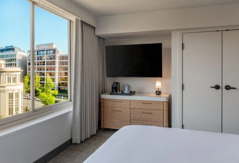 Modern hotel room with a large window, TV, dresser, coffee maker, lamp, and city buildings visible outside in daylight.