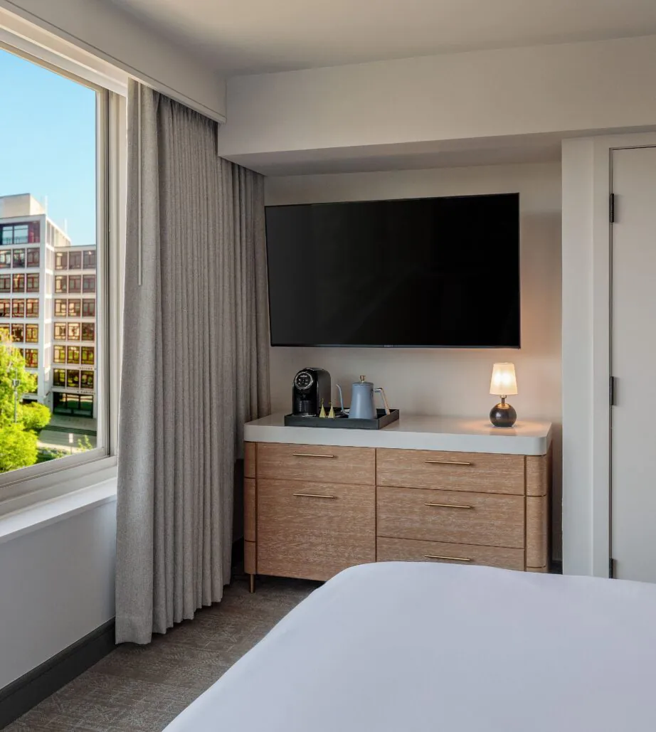 Modern hotel room with a large window, TV, dresser, coffee maker, lamp, and city buildings visible outside in daylight.