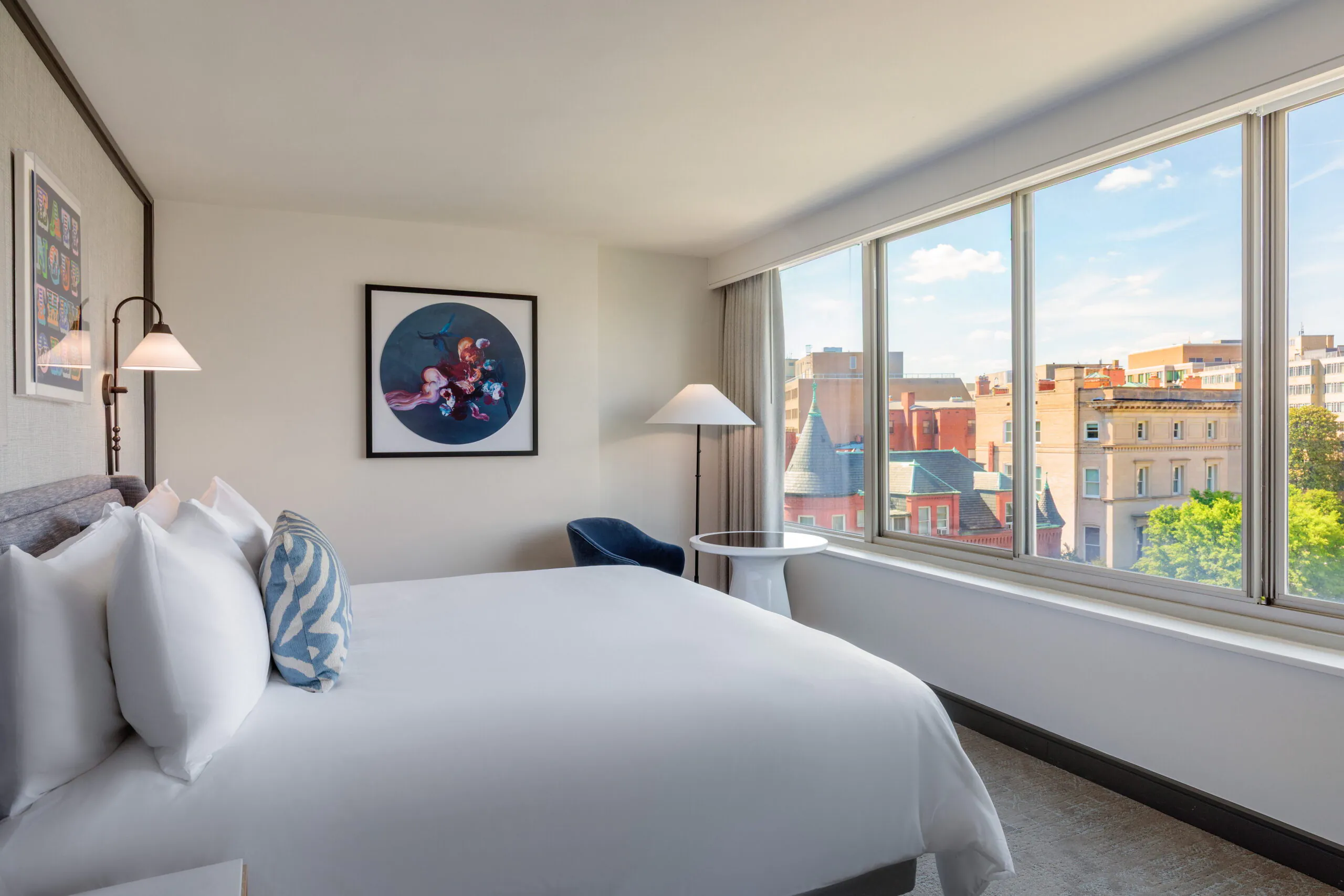 Bright hotel room with a large bed, modern decor, artwork on the wall, and a view of city buildings through wide windows.