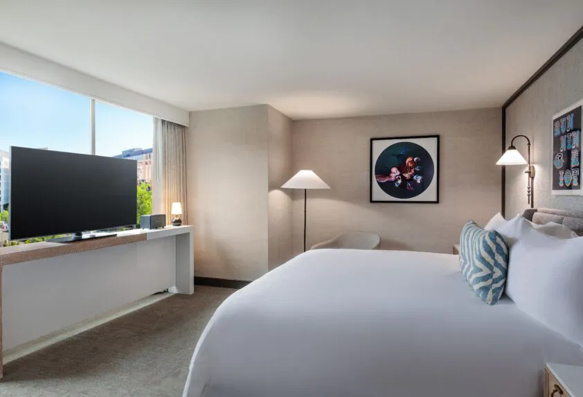 Modern hotel room with a large bed, flat-screen TV, chairs, wall art, and a window letting in natural light with a city view.