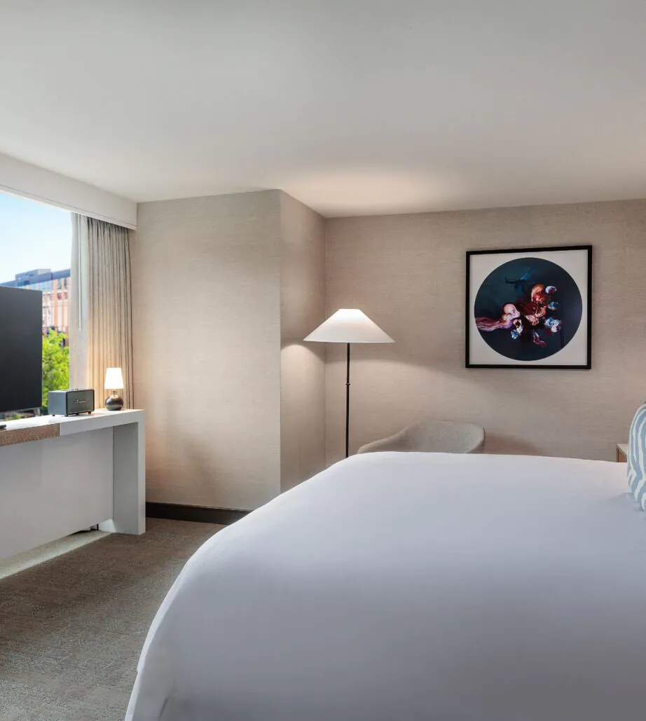 Modern hotel room with a large bed, flat-screen TV, chairs, wall art, and a window letting in natural light with a city view.