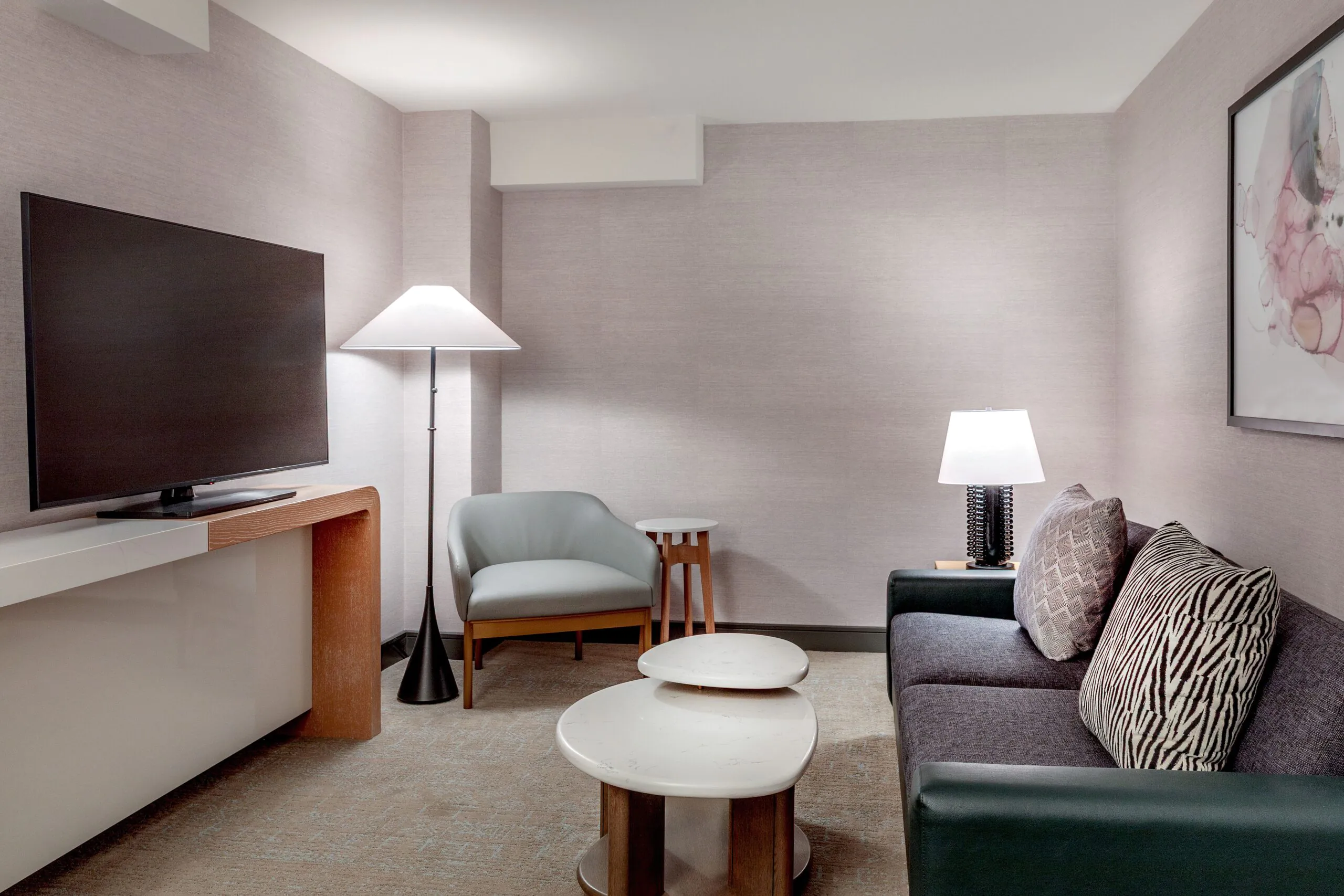 Modern hotel room with a large TV, armchair, sofa, two round coffee tables, floor lamp, table lamp, and wall art on a neutral wall.