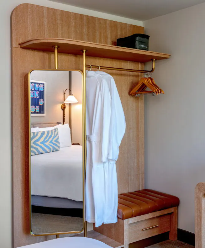 Modern hotel room with an open wardrobe, hanging robe, mirror, wooden bench, coffee maker, and part of a bed and TV visible.