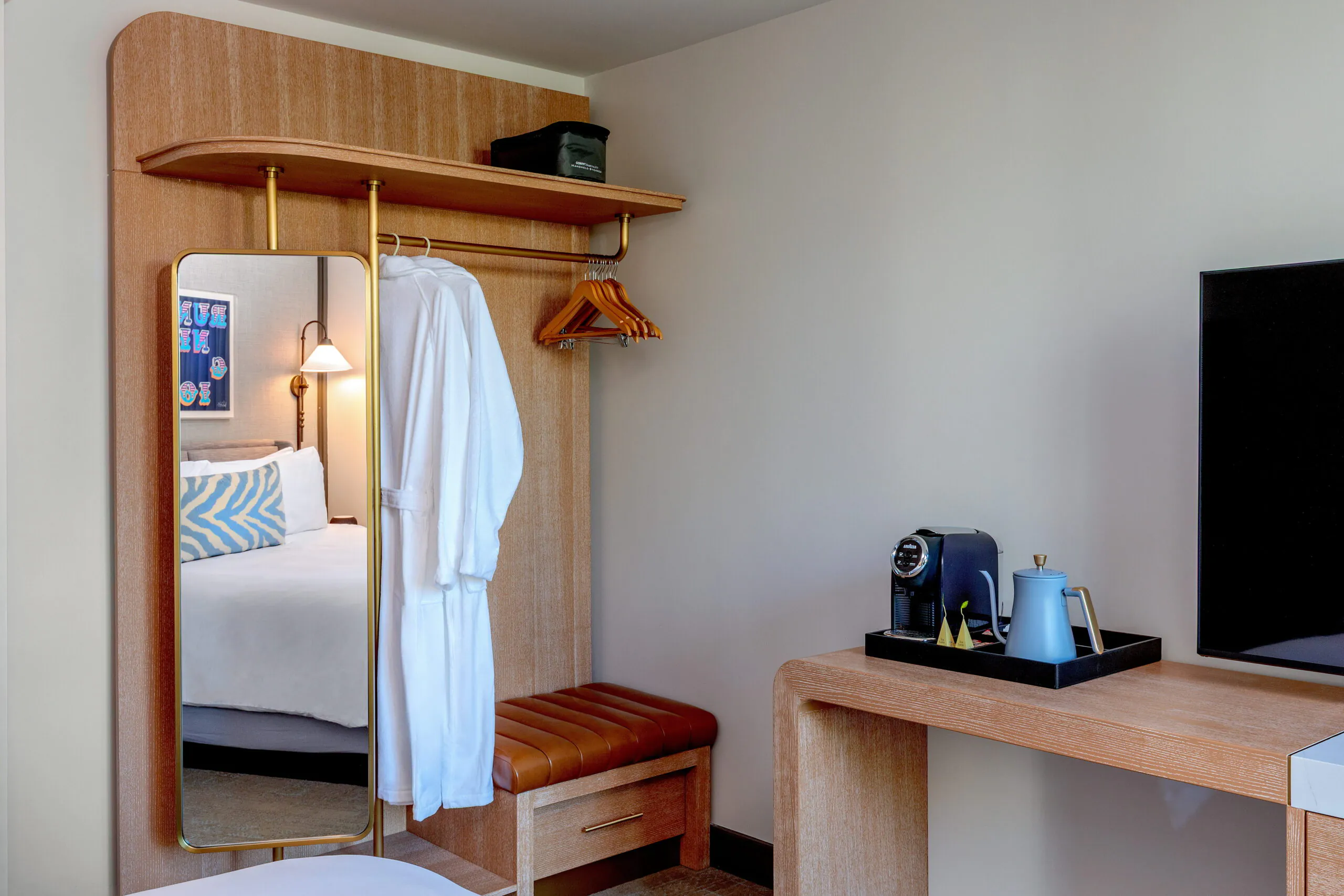 Modern hotel room with an open wardrobe, hanging robe, mirror, wooden bench, coffee maker, and part of a bed and TV visible.