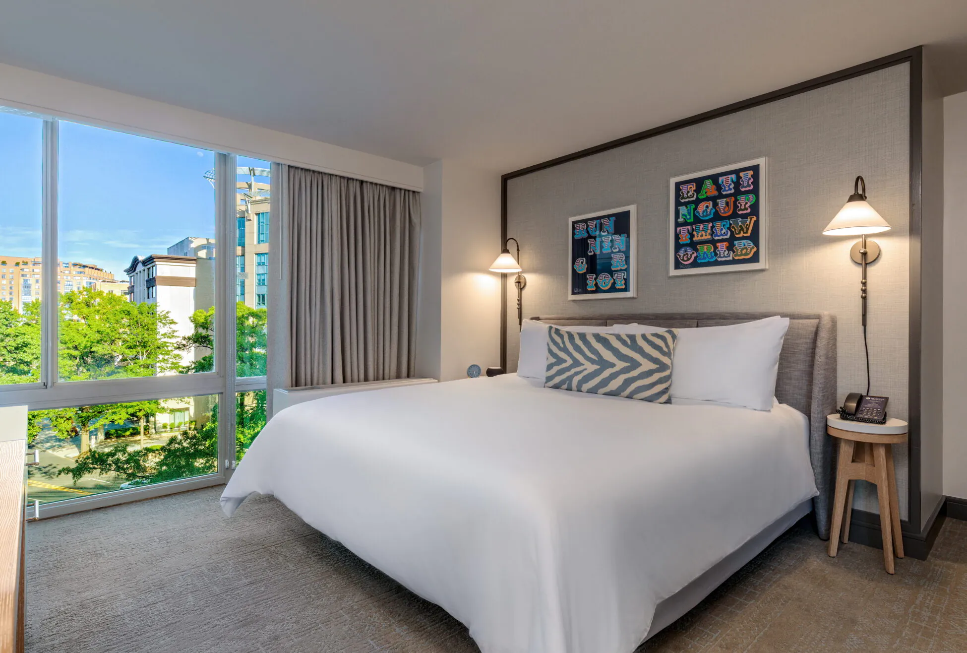 Modern hotel room with a large bed, zebra print pillow, two wall lamps, artwork above the headboard, and a large window with city and trees outside.