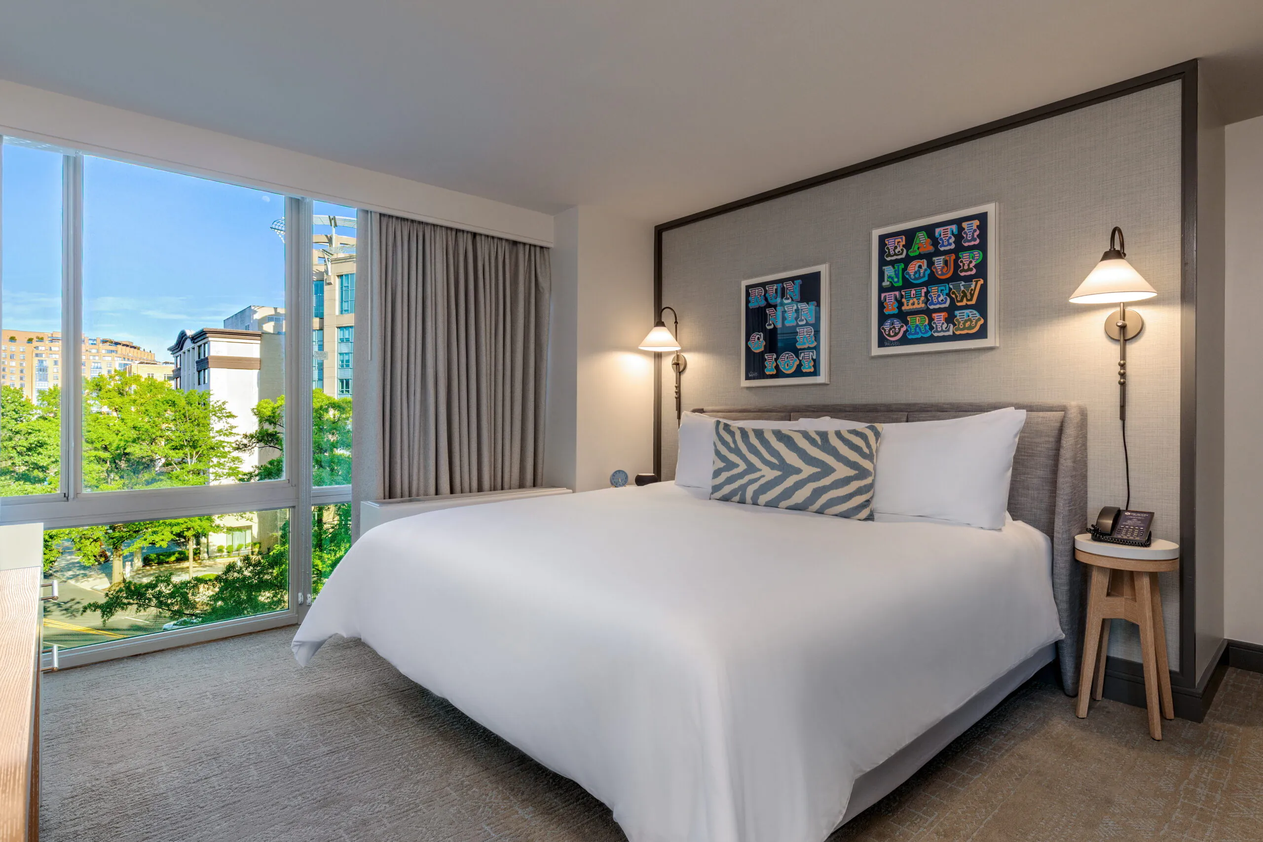 Modern hotel room with a large bed, zebra print pillow, two wall lamps, artwork above the headboard, and a large window with city and trees outside.