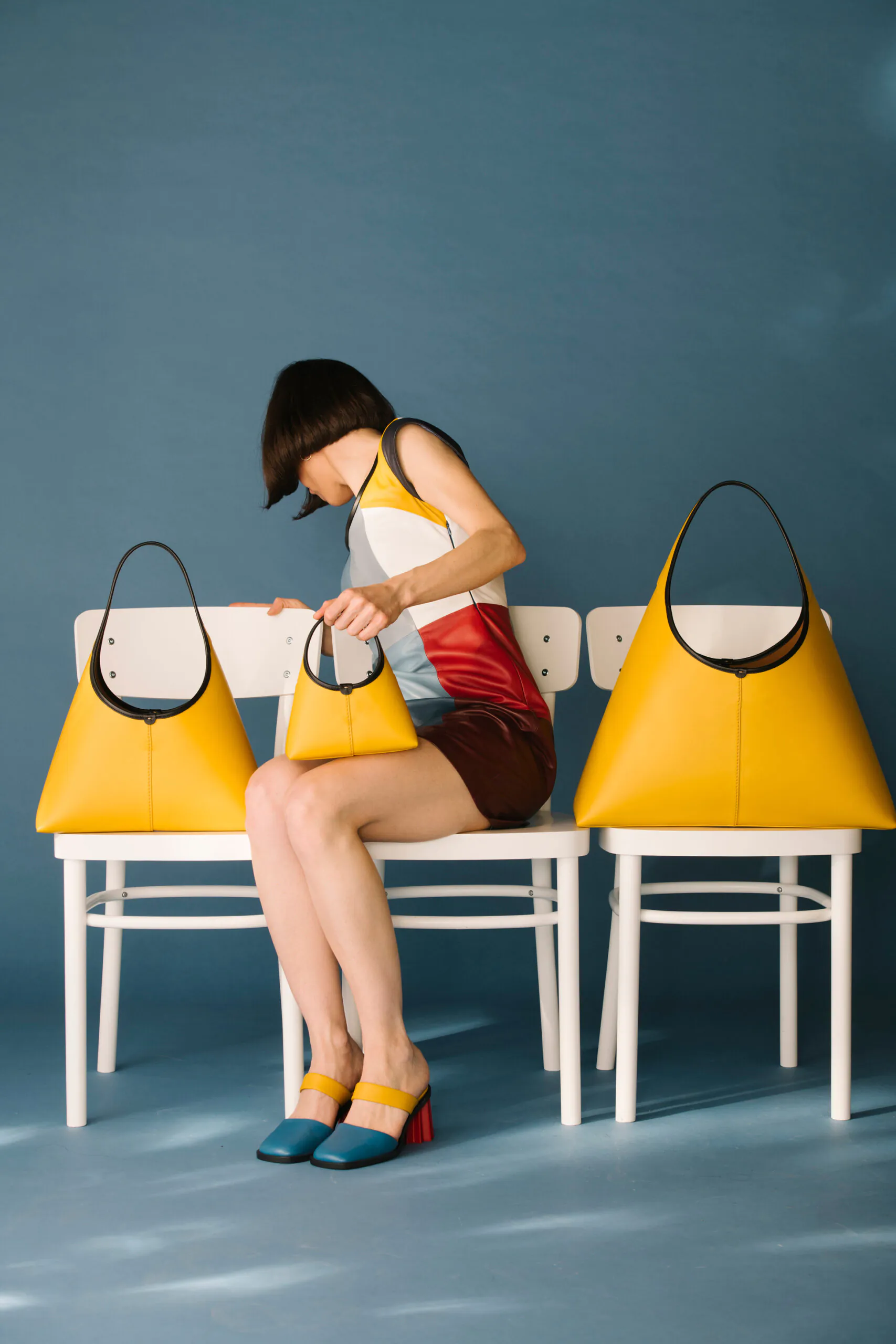 A woman in colorful clothes sits on white chairs with three large yellow handbags, two beside her and one in her lap, against a blue background.
