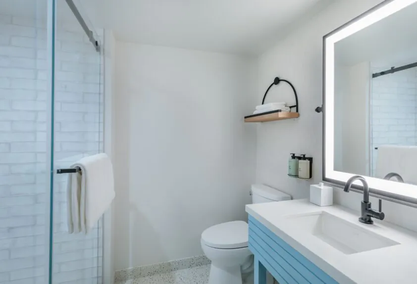 Modern bathroom with a glass shower, toilet, floating shelf with towels, and a sink with a large, illuminated mirror above it.