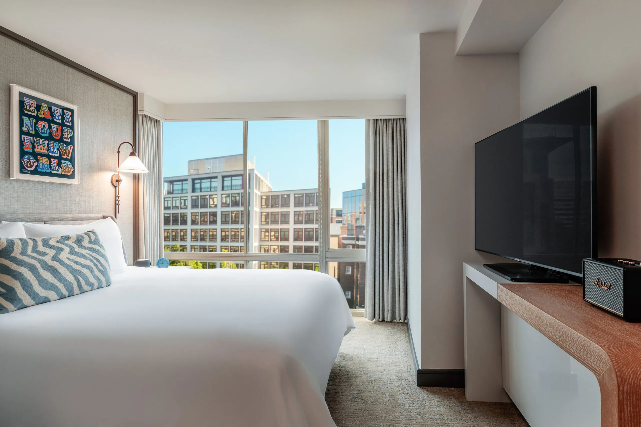 Modern hotel room with a large bed, patterned pillow, wall art, flat-screen TV, and city buildings visible through a large window.