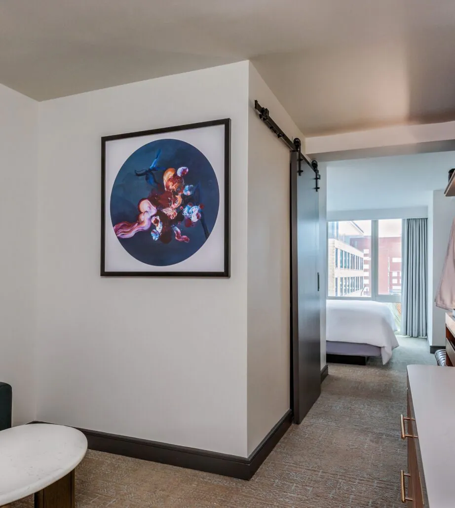 Modern hotel suite with abstract art on the wall, a sitting area, sliding barn door, and a view of a bed in a separate room.
