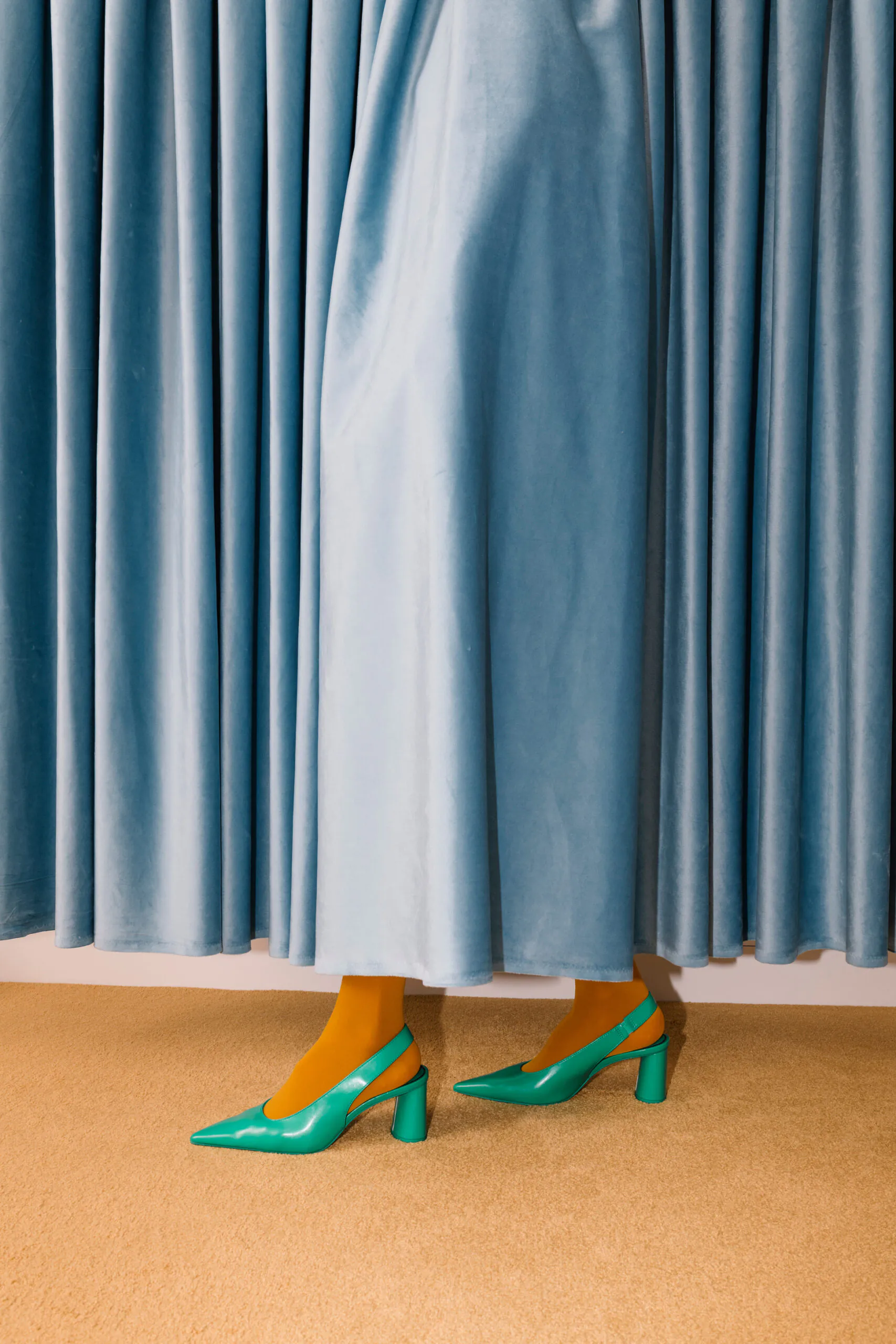 A person in yellow tights and teal heels stands behind a closed light blue curtain, with only their legs and feet visible on a beige carpet.