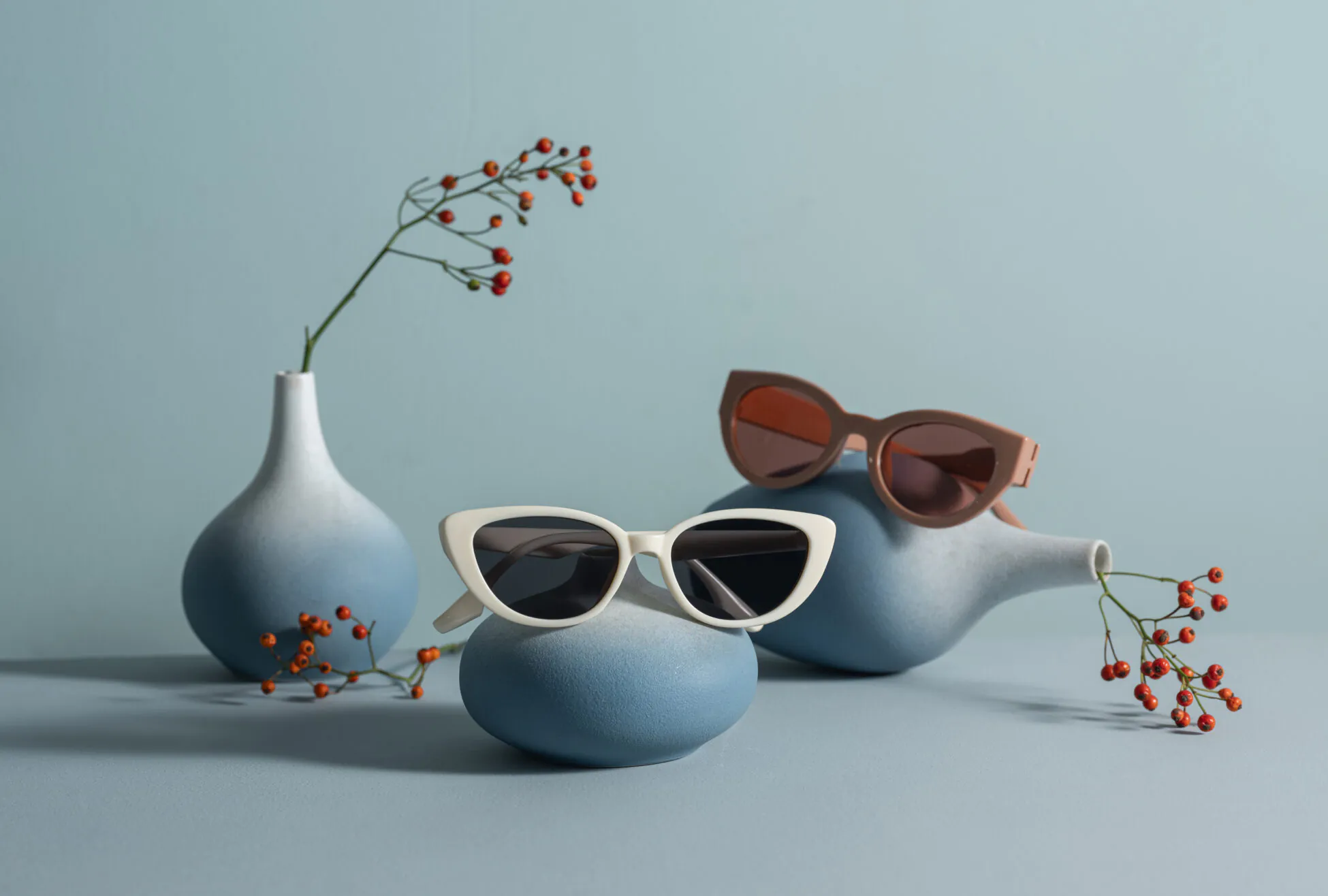 Two pairs of stylish sunglasses rest on blue vases, with small branches of red berries in matching gradient vases, against a light blue background.