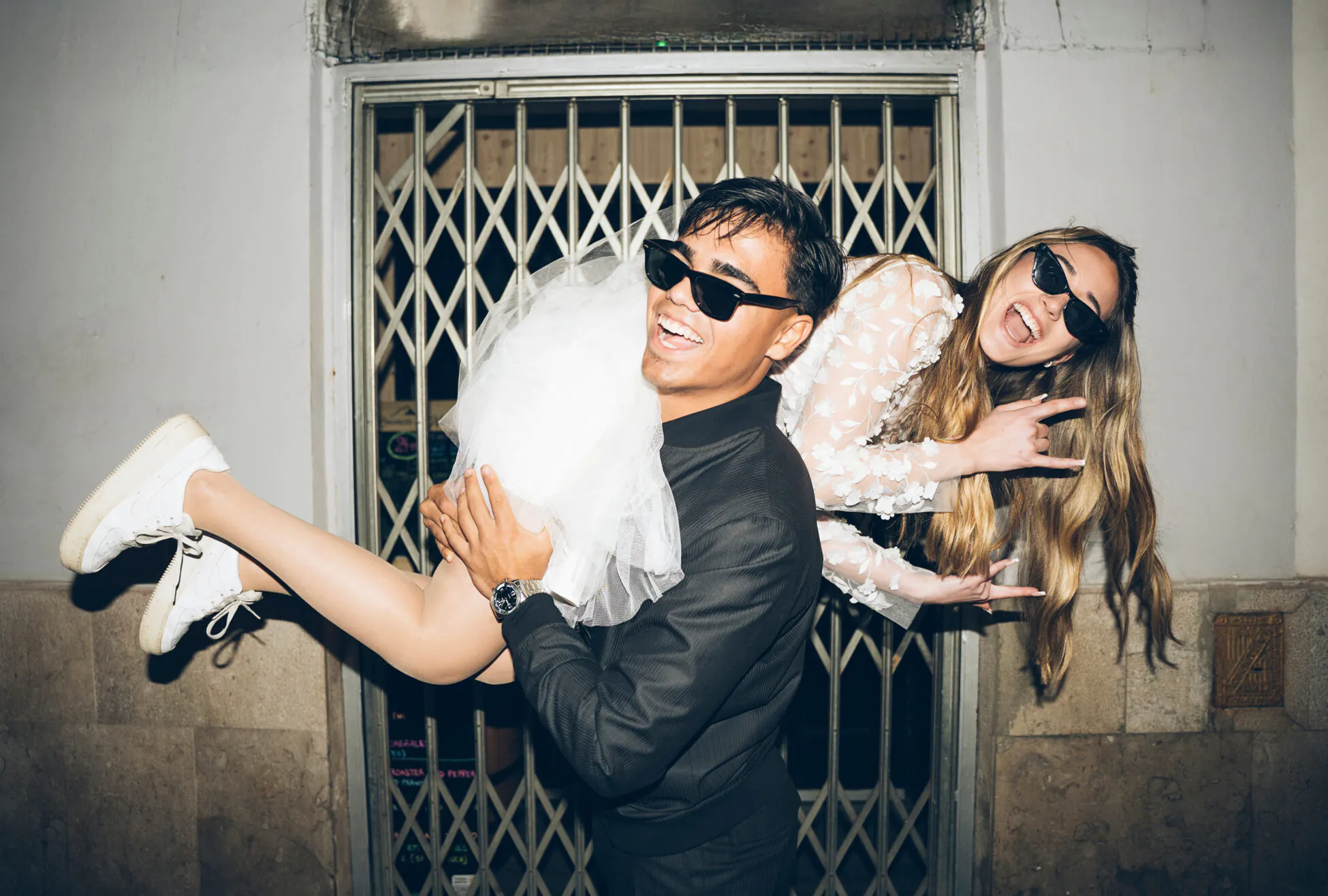 A young man in sunglasses playfully carries a smiling woman in a white dress, also wearing sunglasses, in front of a metal gate.