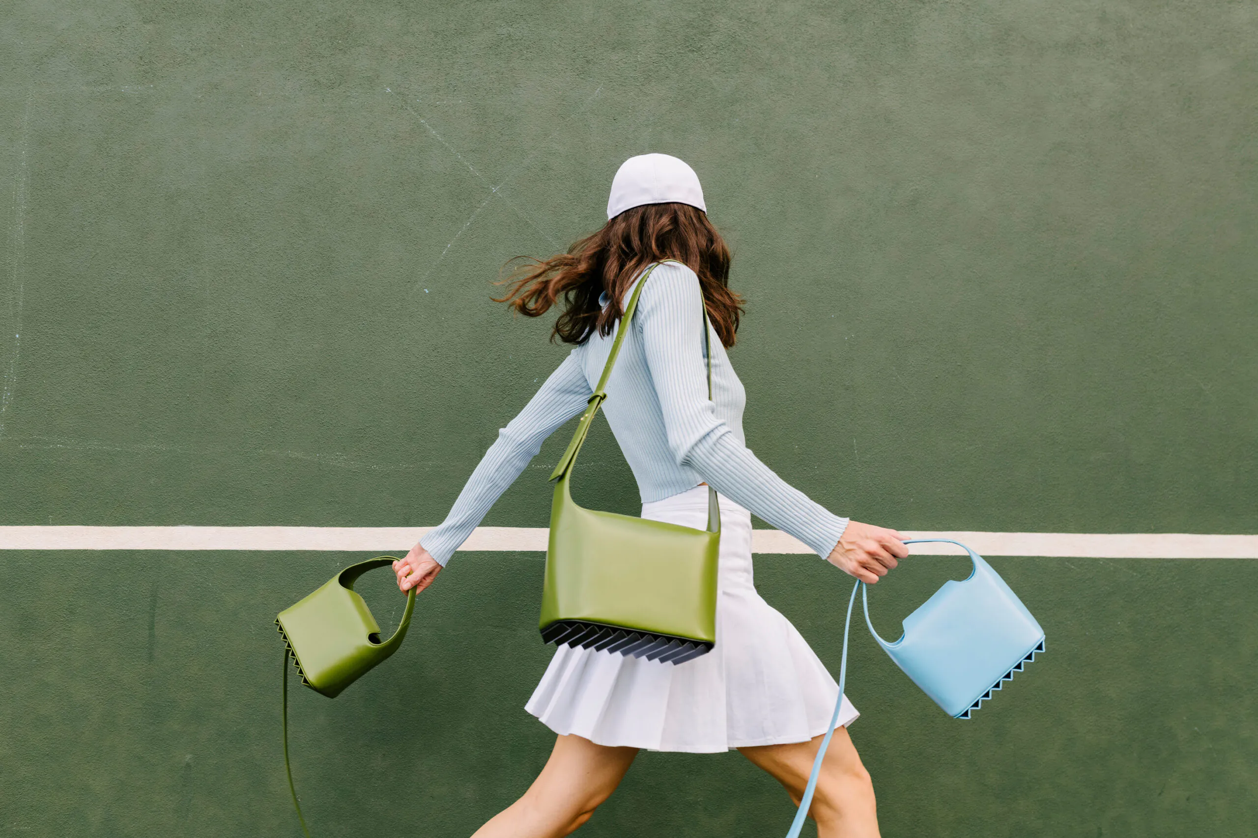 Woman in a white skirt, blue top, and cap walks in front of a green wall, carrying three colorful handbags—green, olive, and blue.