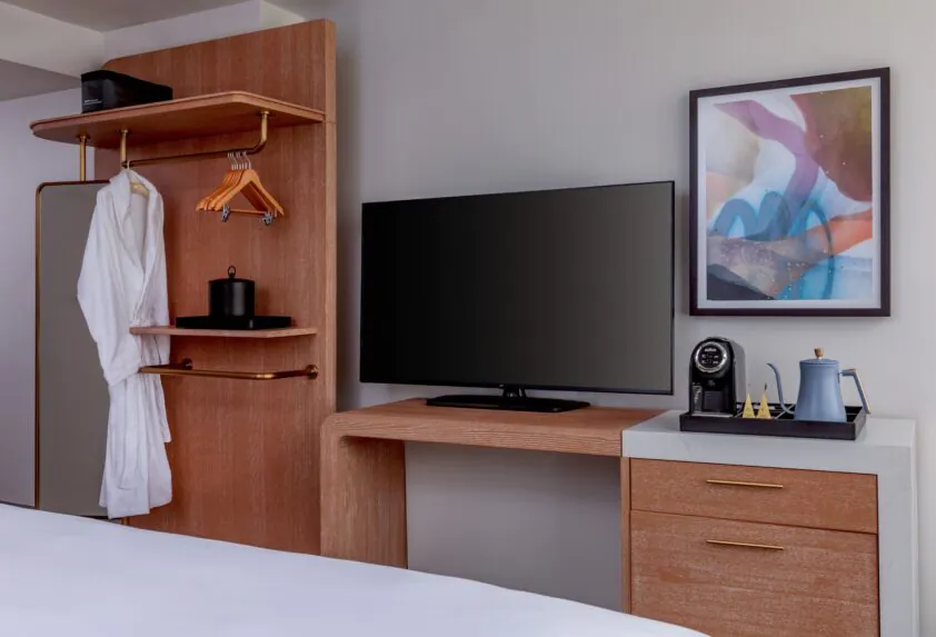 Hotel room with a flat-screen TV, wooden desk, bathrobe, open closet, framed abstract art, and a coffee maker set on a cabinet.