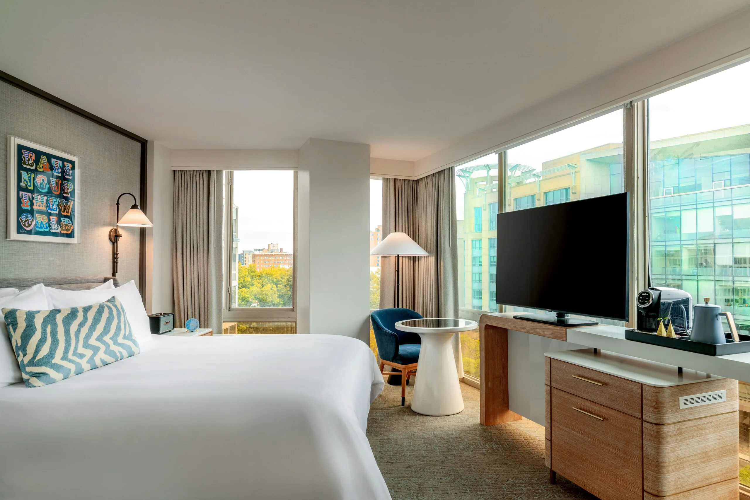 Modern hotel room with a bed, large TV, coffee maker, and floor-to-ceiling windows offering city views and natural light.