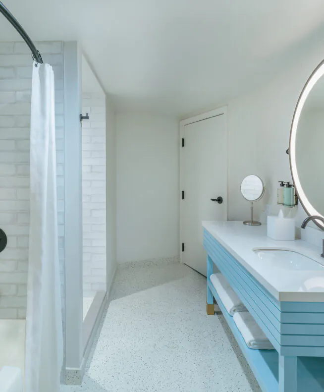 Modern bathroom with a white tub, shower curtain, round backlit mirror, blue vanity with double sinks, and white brick walls.