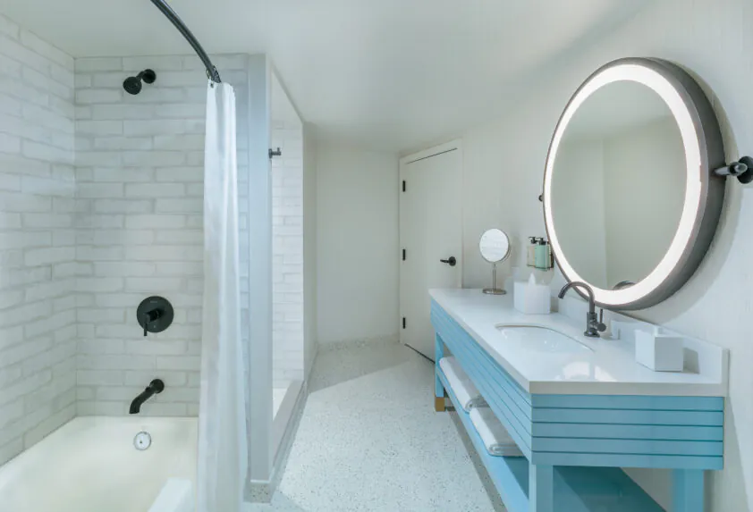 Modern bathroom with a white tub, shower curtain, round backlit mirror, blue vanity with double sinks, and white brick walls.