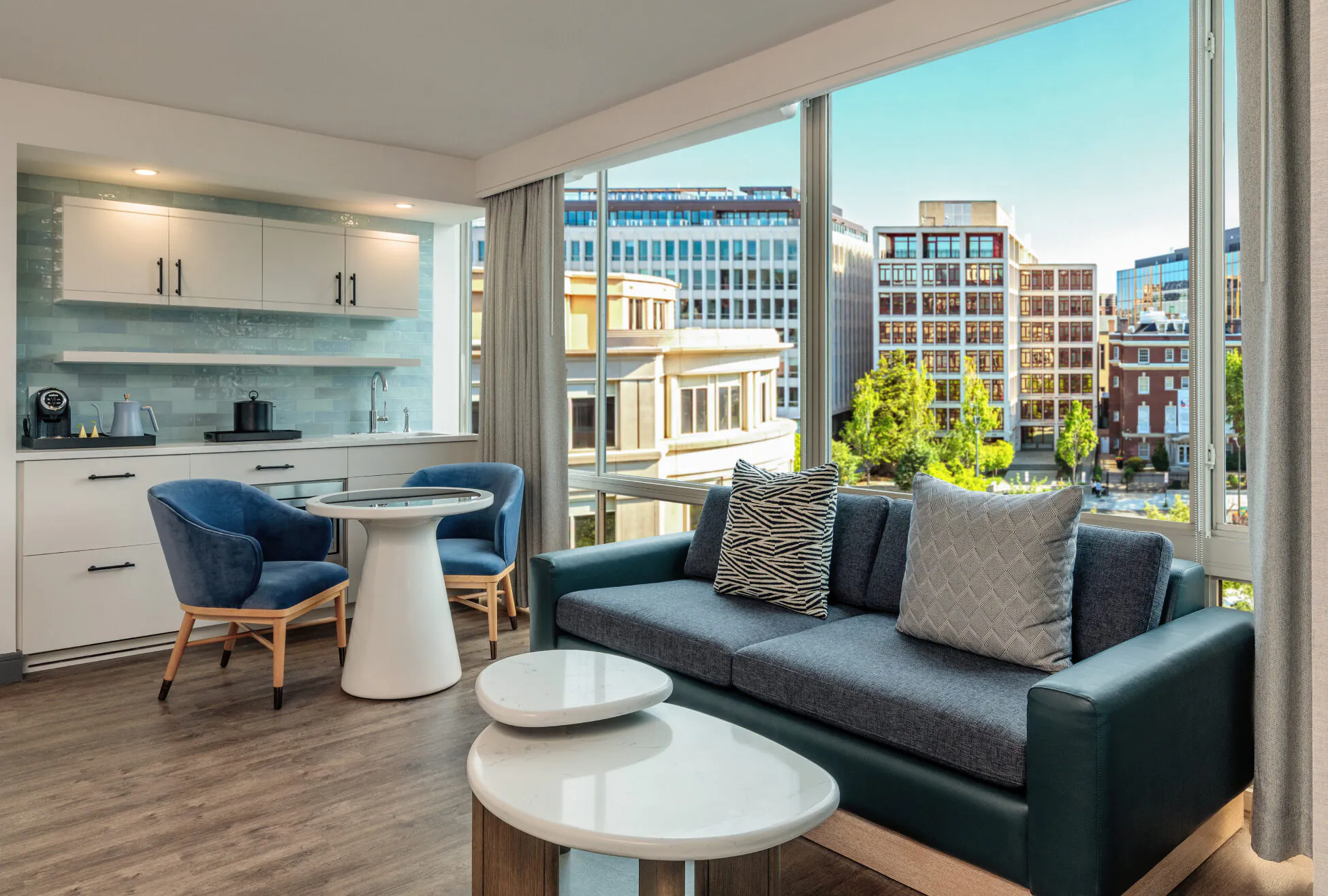 Modern apartment living room with a blue sofa, round tables, two blue chairs, and large windows overlooking city buildings on a sunny day.