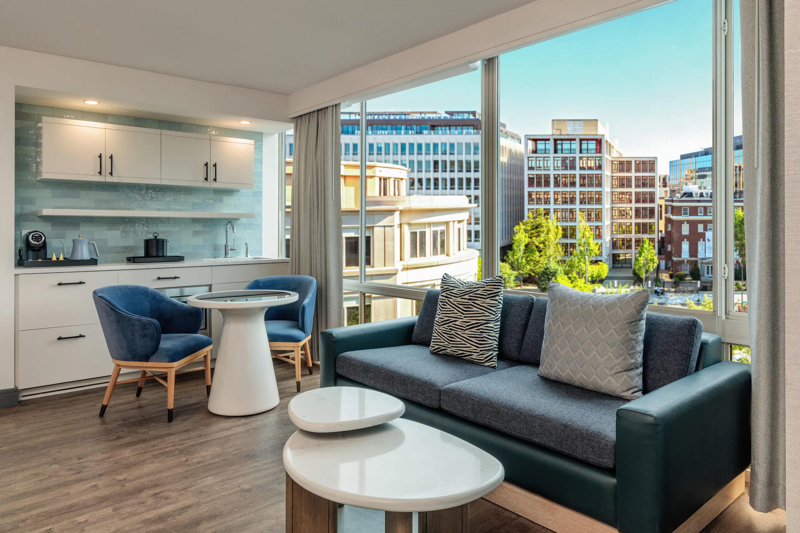 Modern apartment living room with a blue sofa, round tables, two blue chairs, and large windows overlooking city buildings on a sunny day.