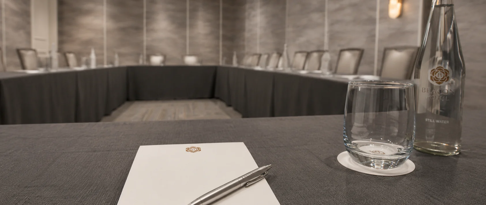 A conference room with a U-shaped table, chairs, a notepad with a pen, a glass of water, and a bottle of water in the foreground.