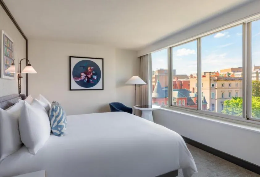 Bright hotel room with a large bed, modern decor, artwork on the wall, and a window offering a city view with buildings and trees.
